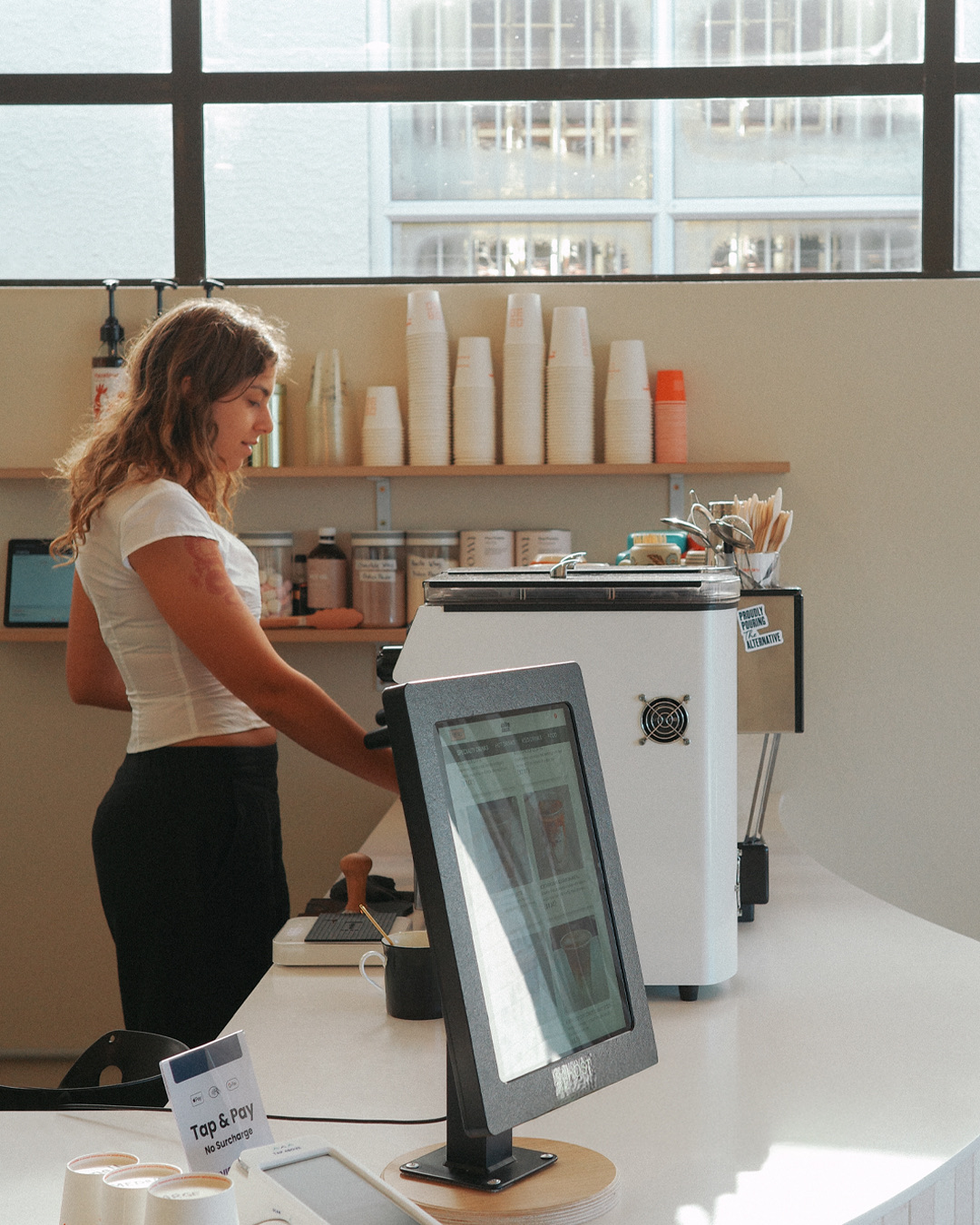 A Daily Dose barista at the cream-tiled counter behind the order tablet.